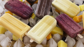 An assortment of colorful popsicles in yellow, purple, and chocolate brown rest atop ice cubes and scattered frozen fruits including mango and berries. The frozen treats are arranged in a visually appealing pattern, creating a refreshing, summery scene.