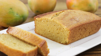 A rectangular loaf of golden-brown mango bread on a white plate, with two slices cut and laid in front. In the background, there are three whole ripe mangoes on a wooden surface.