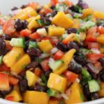 A white bowl filled with a colorful salad featuring mango chunks, black beans, diced red bell peppers, cucumbers, red onions, and chopped cilantro. The ingredients are mixed and displayed closely, highlighting the vibrant, fresh appearance of the dish.