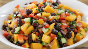 A white bowl filled with a colorful salad featuring mango chunks, black beans, diced red bell peppers, cucumbers, red onions, and chopped cilantro. The ingredients are mixed and displayed closely, highlighting the vibrant, fresh appearance of the dish.