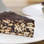 A slice of chocolate biscuit cake is placed on a white plate. The cake is made up of a dense mixture of chocolate and crumbled biscuits, with a smooth, shiny chocolate layer on top. There is another similar slice visible in the background.