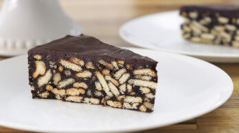 A slice of chocolate biscuit cake is placed on a white plate. The cake is made up of a dense mixture of chocolate and crumbled biscuits, with a smooth, shiny chocolate layer on top. There is another similar slice visible in the background.