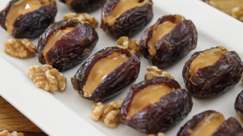 A white rectangular plate holds several dates stuffed with a creamy filling. Nuts are scattered around the dates, adding texture and contrast to the dish.