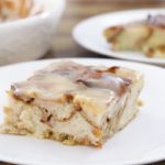 A close-up of a square serving of glazed cinnamon roll casserole on a white plate. The dish has layers of cinnamon swirls topped with a glossy glaze. Another plate with the same casserole is blurred in the background, along with a large serving dish.