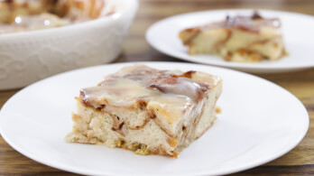 A close-up of a square serving of glazed cinnamon roll casserole on a white plate. The dish has layers of cinnamon swirls topped with a glossy glaze. Another plate with the same casserole is blurred in the background, along with a large serving dish.