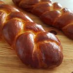 Two freshly baked challah breads are on a round wooden board. The loaves have a golden-brown, glossy crust and are intricately braided. The board rests on a light-colored surface.