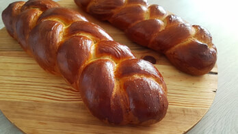 Two freshly baked challah breads are on a round wooden board. The loaves have a golden-brown, glossy crust and are intricately braided. The board rests on a light-colored surface.