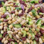 A vibrant salad in a bowl, featuring kidney beans, cannellini beans, chickpeas, sliced red onions, diced cucumbers, and finely chopped herbs, all mixed together, resting on a wooden surface.