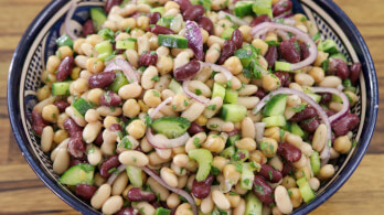 A vibrant salad in a bowl, featuring kidney beans, cannellini beans, chickpeas, sliced red onions, diced cucumbers, and finely chopped herbs, all mixed together, resting on a wooden surface.