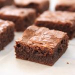 A close-up view of several chocolate brownies arranged on parchment paper. The brownies are cut into neat squares, with a rich, fudgy texture and a slightly cracked top, indicating a perfect bake.