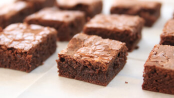 A close-up view of several chocolate brownies arranged on parchment paper. The brownies are cut into neat squares, with a rich, fudgy texture and a slightly cracked top, indicating a perfect bake.