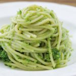 A neatly arranged serving of spaghetti with pesto sauce is presented on a white plate. The pasta is coated evenly with green pesto, and the background is a blurred wooden surface.