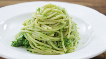 A neatly arranged serving of spaghetti with pesto sauce is presented on a white plate. The pasta is coated evenly with green pesto, and the background is a blurred wooden surface.