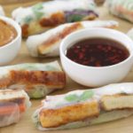 A wooden board displaying several fresh spring rolls, each filled with tofu, vegetables, and herbs wrapped in translucent rice paper. Two small bowls of dipping sauces, one with a brown peanut sauce and the other with a red chili sauce, are placed on the board.