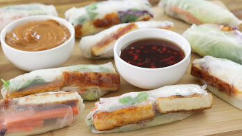 A wooden board displaying several fresh spring rolls, each filled with tofu, vegetables, and herbs wrapped in translucent rice paper. Two small bowls of dipping sauces, one with a brown peanut sauce and the other with a red chili sauce, are placed on the board.