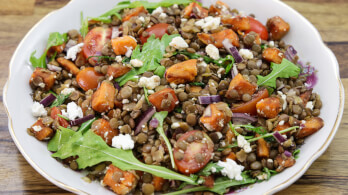 A colorful salad in a white bowl features a mix of lentils, arugula, cherry tomatoes, roasted sweet potatoes, red onions, and crumbled feta cheese. The ingredients are tossed together, creating a vibrant and healthy dish.