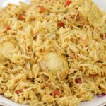 A plate of spicy biryani rice, featuring a mix of yellow rice, chunks of vegetables, and two boiled eggs nestled within. The dish is served on a white plate with a wooden background.