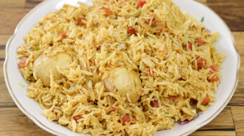 A plate of spicy biryani rice, featuring a mix of yellow rice, chunks of vegetables, and two boiled eggs nestled within. The dish is served on a white plate with a wooden background.