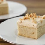 A close-up of a vanilla cake square on a white plate, topped with light frosting and chopped nuts. Another plate with a similar cake is blurred in the background, all set on a wooden surface.