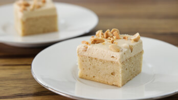 A close-up of a vanilla cake square on a white plate, topped with light frosting and chopped nuts. Another plate with a similar cake is blurred in the background, all set on a wooden surface.