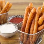Two metal baskets filled with crunchy, golden-brown fried mozzarella sticks sit on a wooden table. One small bowl of marinara sauce and another small bowl of aioli dip are placed between the baskets.