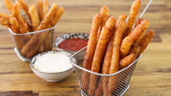 Two metal baskets filled with crunchy, golden-brown fried mozzarella sticks sit on a wooden table. One small bowl of marinara sauce and another small bowl of aioli dip are placed between the baskets.