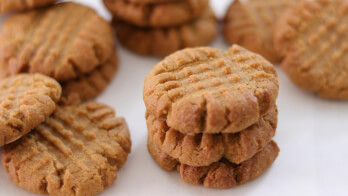 A close-up of several peanut butter cookies stacked and arranged on a white surface. The cookies are golden-brown with criss-cross fork marks on top, indicating they are freshly baked.