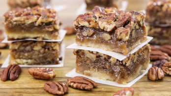 A close-up of two stacked pecan pie bars on a wooden surface. The bars feature a crumbly crust, a gooey caramel-like filling, and a generous topping of pecans. Additional pecan pie bars and loose pecan nuts are scattered in the background.