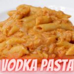 A white plate filled with penne pasta in a creamy tomato vodka sauce. The text "VODKA PASTA" is prominently displayed at the bottom of the image in bold, pink letters.