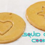 Two round cookies rest on a white surface. One cookie has a heart shape pressed into it, and the other has a star shape. The text "SQUID GAME COOKIE" appears in the bottom right corner.