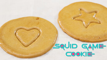 Two round cookies rest on a white surface. One cookie has a heart shape pressed into it, and the other has a star shape. The text "SQUID GAME COOKIE" appears in the bottom right corner.