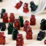 A variety of gummy bear candies in red, green, and black are scattered on a wooden surface. Some gummies are standing upright while others are lying down. A glass jar is tipped over, with more candies spilling out of it.
