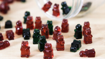 A variety of gummy bear candies in red, green, and black are scattered on a wooden surface. Some gummies are standing upright while others are lying down. A glass jar is tipped over, with more candies spilling out of it.