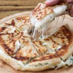 A close-up of a cheese-stuffed, sliced flatbread on a wooden plate. A piece is being lifted, showing melted cheese stretching from the flatbread. The flatbread has a golden-brown, crispy crust. The background is a wooden surface.
