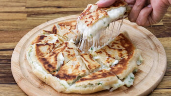 A close-up of a cheese-stuffed, sliced flatbread on a wooden plate. A piece is being lifted, showing melted cheese stretching from the flatbread. The flatbread has a golden-brown, crispy crust. The background is a wooden surface.