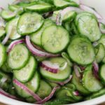 A white bowl filled with a cucumber salad consisting of thinly sliced cucumbers, red onion slivers, and dill. The ingredients are lightly coated with a dressing, creating a fresh and vibrant appearance. The background is a wooden surface.