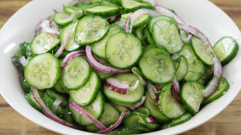 A white bowl filled with a cucumber salad consisting of thinly sliced cucumbers, red onion slivers, and dill. The ingredients are lightly coated with a dressing, creating a fresh and vibrant appearance. The background is a wooden surface.