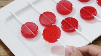 Seven red, round lollipops arranged in a grid pattern on a white rectangular plate. A hand is holding the stick of one lollipop at the bottom right of the image.