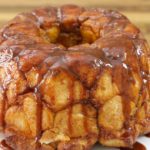 A glazed, golden-brown monkey bread is displayed on a white plate, resting on a wooden surface. The bread appears moist and sticky, with caramelized sauce dripping over the soft, pull-apart pieces.