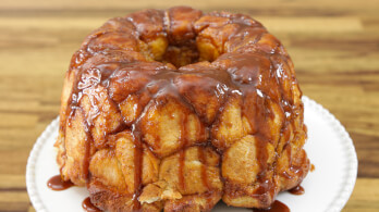 A glazed, golden-brown monkey bread is displayed on a white plate, resting on a wooden surface. The bread appears moist and sticky, with caramelized sauce dripping over the soft, pull-apart pieces.