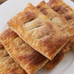 A close-up of a plate topped with four golden-brown rectangular pastries. The pastries have a flaky texture and are arranged in a neat stack. The plate is placed on a wooden surface, enhancing the warm, inviting appearance of the pastries.