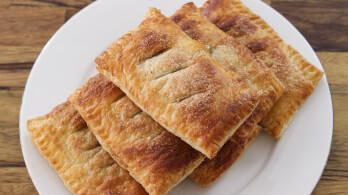 A close-up of a plate topped with four golden-brown rectangular pastries. The pastries have a flaky texture and are arranged in a neat stack. The plate is placed on a wooden surface, enhancing the warm, inviting appearance of the pastries.