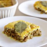 Two white plates each hold a serving of bobotie, a South African dish made of spiced minced meat topped with an egg-based custard. A bay leaf decorates the top layer of the portion closest to the camera. In the background is a white baking dish with more bobotie.