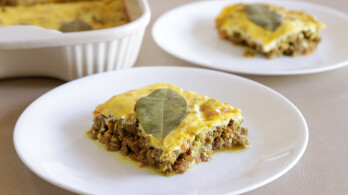 Two white plates each hold a serving of bobotie, a South African dish made of spiced minced meat topped with an egg-based custard. A bay leaf decorates the top layer of the portion closest to the camera. In the background is a white baking dish with more bobotie.