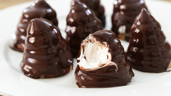 A plate of chocolate-coated marshmallow treats is arranged, with one treat partially bitten to reveal the fluffy white marshmallow filling inside. The conical treats have a glossy, smooth chocolate exterior.