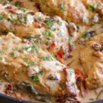 A skillet filled with creamy Tuscan chicken. The dish features browned chicken breasts cooked in a rich, creamy sauce with visible ingredients like sun-dried tomatoes, spinach, and herbs. The sauce is thick and garnished with freshly chopped parsley.