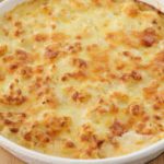 A baking dish filled with golden-brown macaroni and cheese, featuring a bubbly, crispy top layer of melted cheese. The dish rests on a wooden surface, showcasing its creamy and rich texture. Handles are visible on each side of the dish.