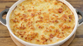 A baking dish filled with golden-brown macaroni and cheese, featuring a bubbly, crispy top layer of melted cheese. The dish rests on a wooden surface, showcasing its creamy and rich texture. Handles are visible on each side of the dish.