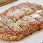 A rectangular, cheesy garlic bread topped with melted cheese is cut into evenly-sized sticks and arranged on parchment paper, set on a wooden surface. The bread is golden brown with visible herbs and a dusting of grated Parmesan cheese.