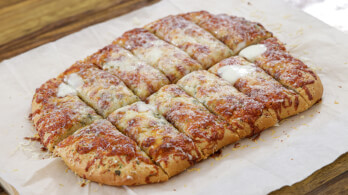 A rectangular, cheesy garlic bread topped with melted cheese is cut into evenly-sized sticks and arranged on parchment paper, set on a wooden surface. The bread is golden brown with visible herbs and a dusting of grated Parmesan cheese.